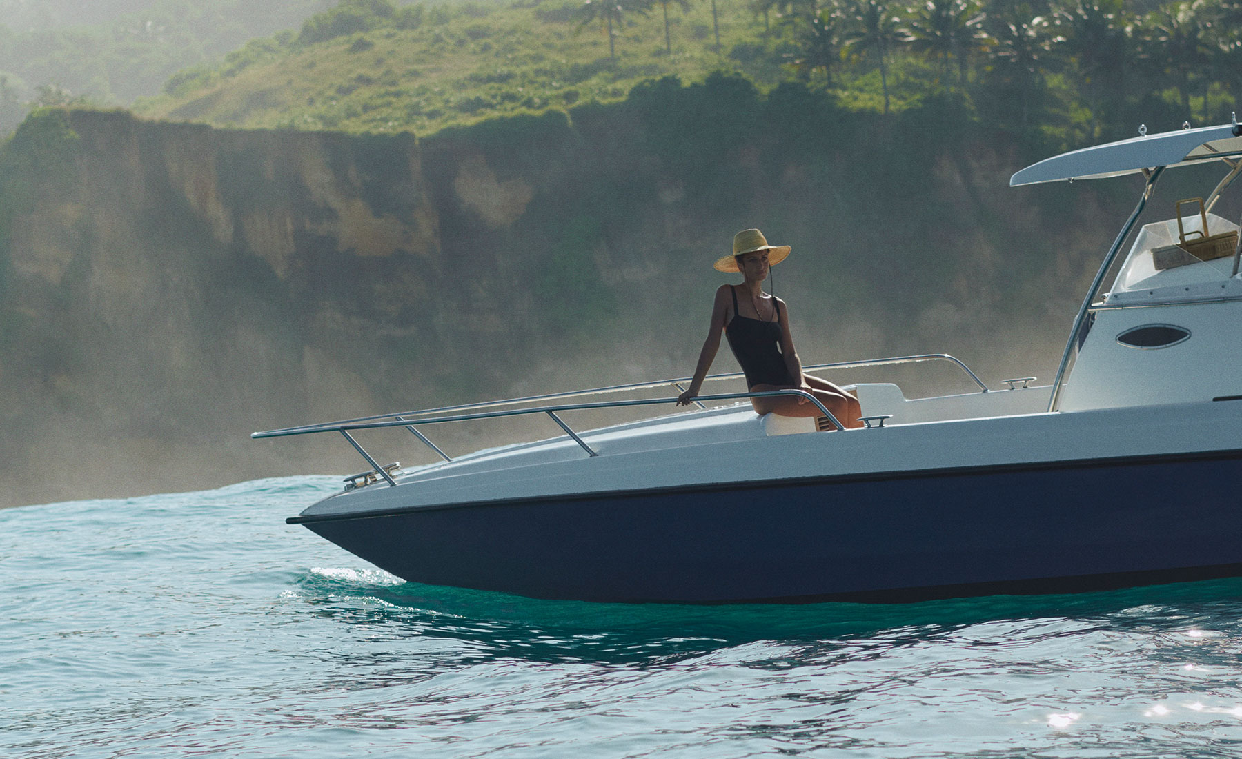 A woman in a sunhat and black swimsuit relaxes at the bow of a luxury speedboat off the coast of Sumba, with dramatic cliffs and lush greenery in the background—capturing the spirit of ocean exploration at NIHI Sumba.