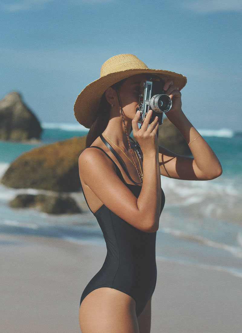 Guest in a straw hat photographing Nihiwatu Beach with a vintage camera at NIHI Sumba, capturing the island’s wild beauty and coastal charm.