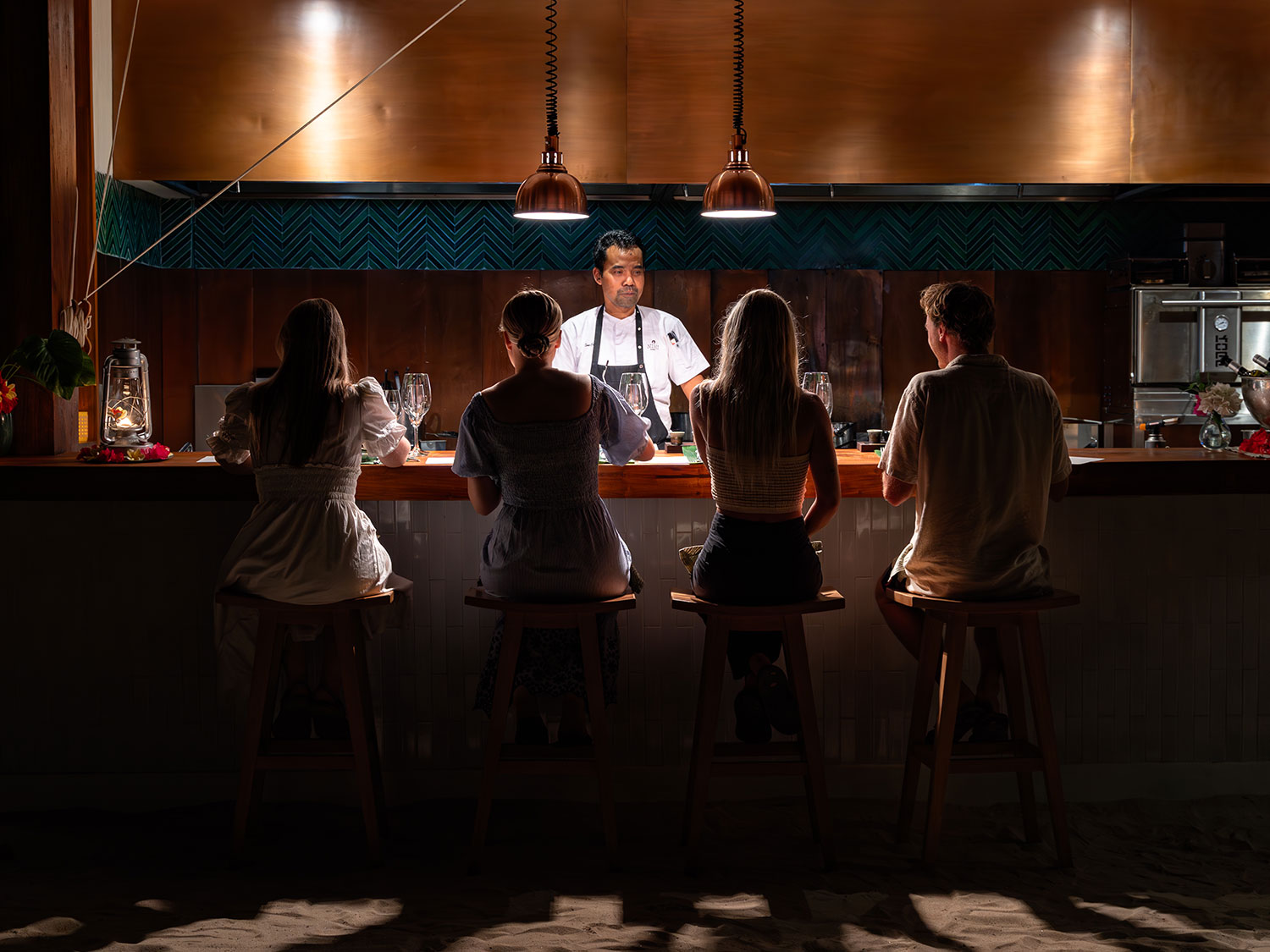 A group of guests seated at the Chef’s Table at Nio, immersed in the intimate dining experience as the chef stands behind the counter. The setting is enhanced by warm lighting and elegant decor, reflecting NIHI Sumba’s signature fusion of East and West cuisines.