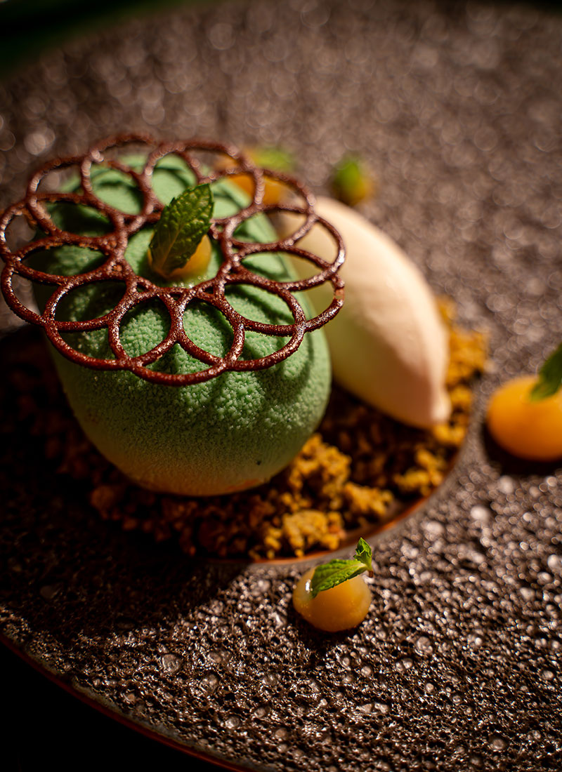 An artfully presented dessert at the Chef's Table at Nio, featuring green mousse with a delicate chocolate lattice, surrounded by mint leaves and vibrant garnishes, showcasing the fine culinary craftsmanship of the dining experience.