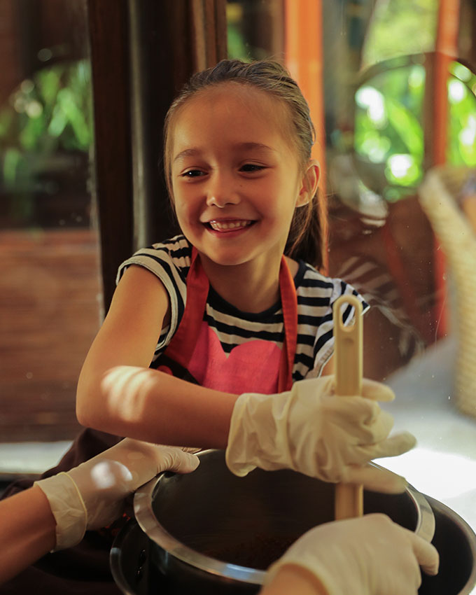 Smiling young girl in an apron and gloves stirring melted chocolate during a hands-on kids’ chocolate-making class at NIHI Sumba’s Chris & Charly’s chocolate factory.
