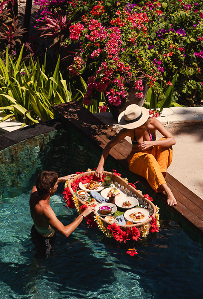 Couple enjoying a tropical floating breakfast in a private pool surrounded by vibrant flowers and lush greenery at NIHI Sumba.