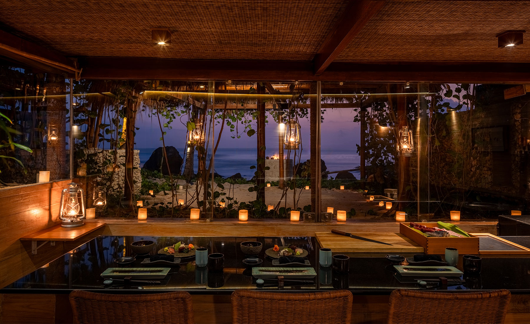 Intimate evening setting at Kaboku, NIHI Sumba’s Japanese omakase restaurant, featuring a candlelit table with a view of the beach and ocean framed by lush foliage and softly glowing lanterns.