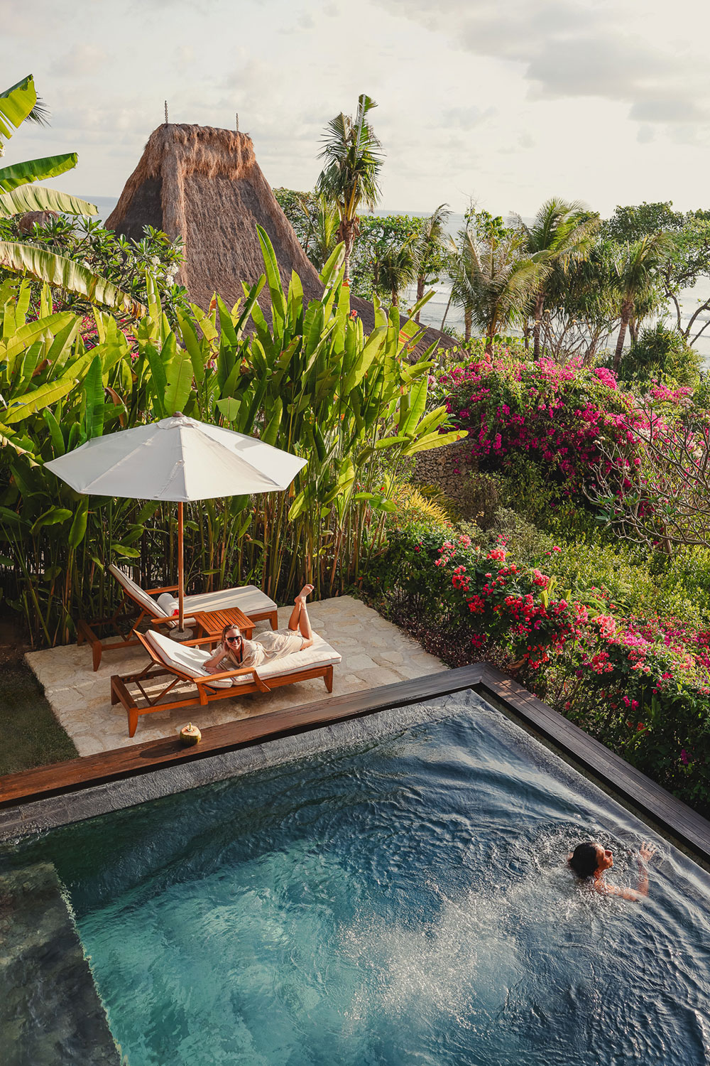 Guests relaxing at the private pool of the Kanatar Villa at NIHI® Sumba, with one person lounging under an umbrella and another swimming, surrounded by tropical plants and vibrant flowers.