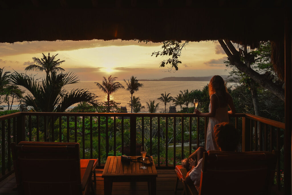 Guests enjoying a sunset view over the Indian Ocean from the private balcony of Kanatar villa at NIHI Sumba, surrounded by tropical palms and tranquil luxury.