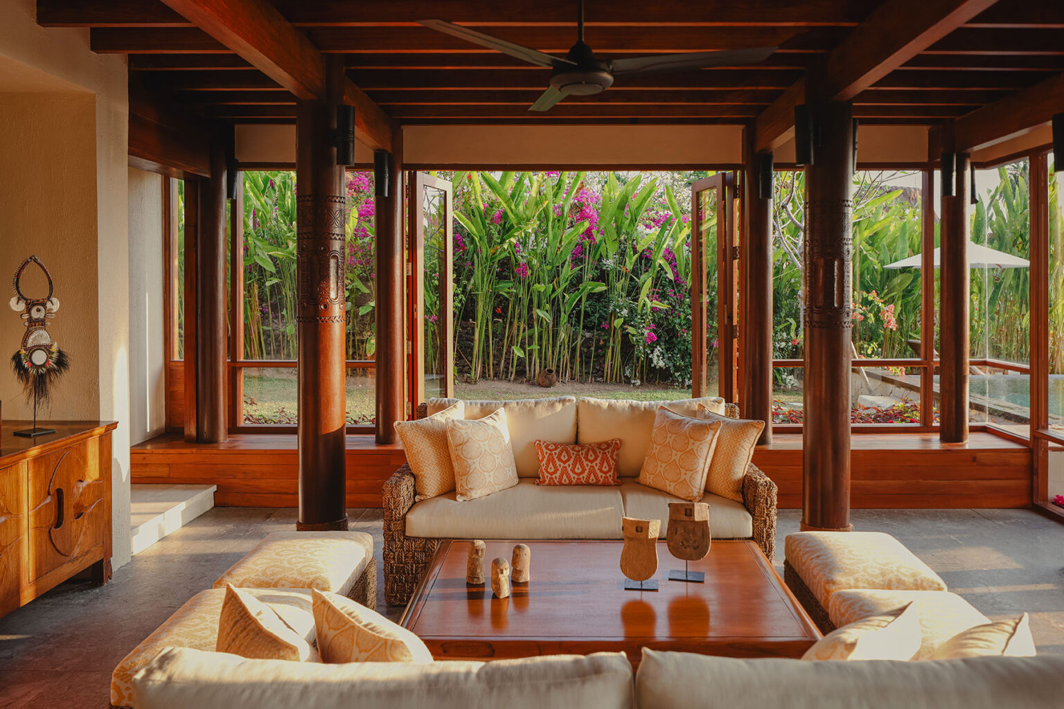 Sunlit indoor lounge at Kanatar villa in NIHI Sumba, featuring a cozy rattan sofa set with views of lush tropical foliage and blooming flowers just beyond the open windows.