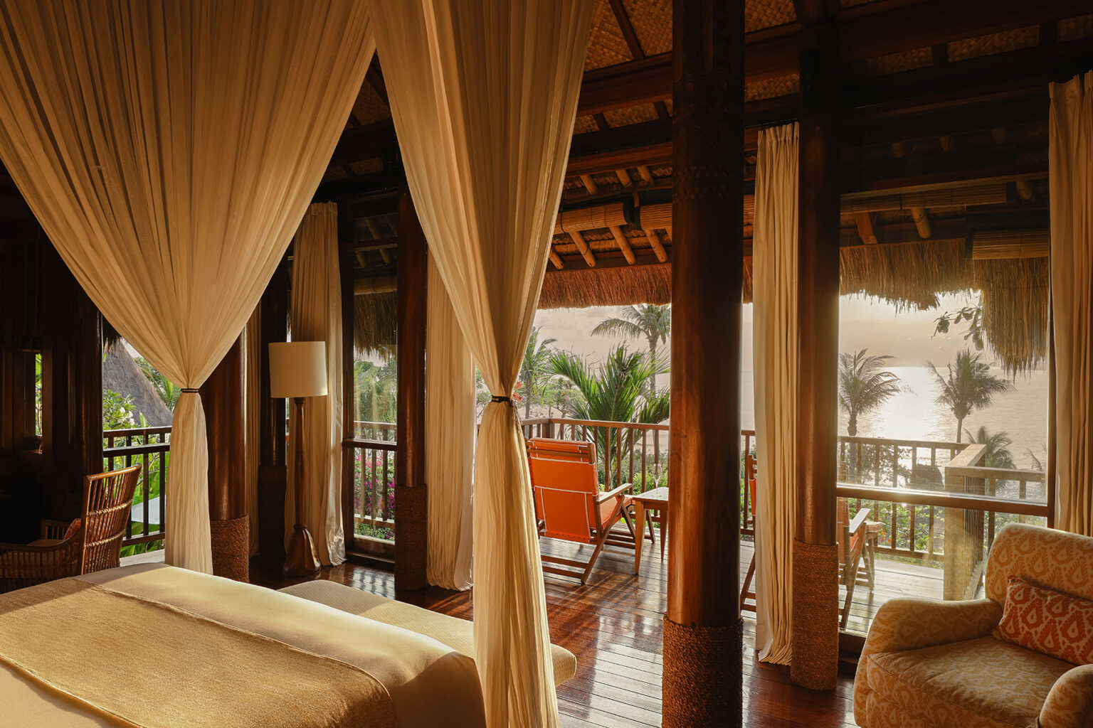 Elegant canopy bed in a sunlit bedroom at NIHI Sumba’s Kanatar villa, featuring rich wooden interiors, flowing curtains, and a private balcony overlooking tropical palm trees and an ocean view.