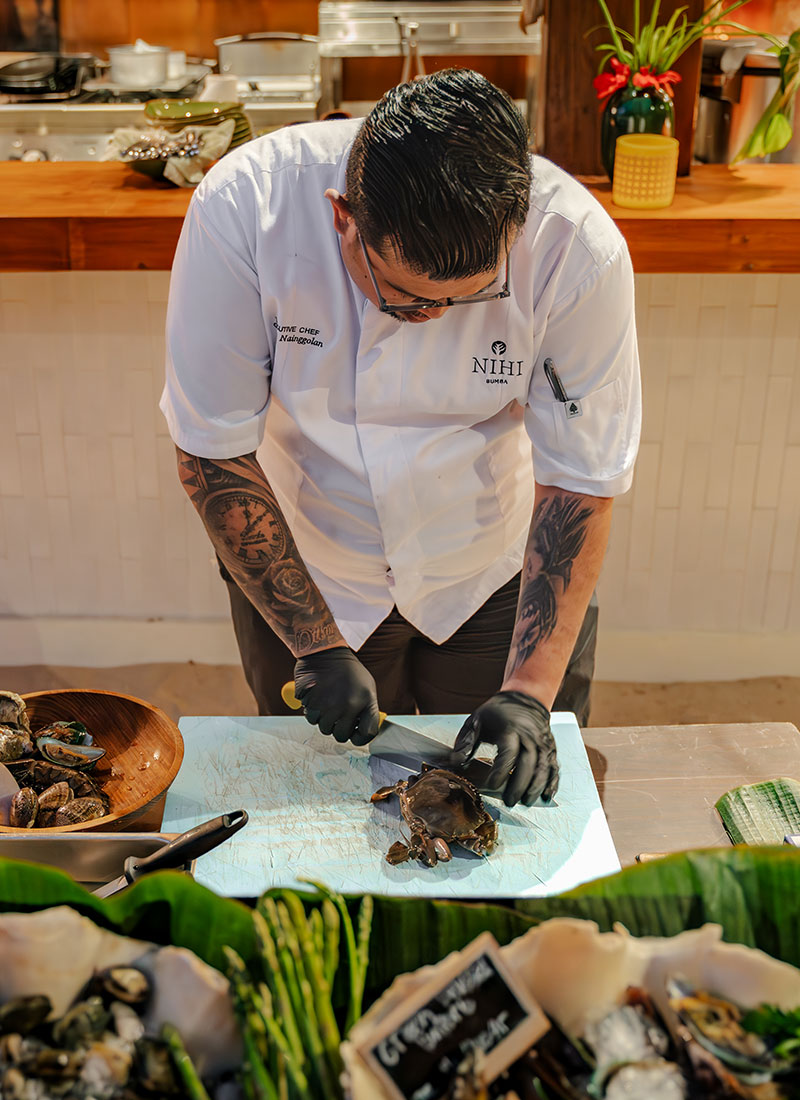 A chef at NIHI Sumba skillfully preparing crab at the Seafood Market. Dressed in a NIHI-branded chef jacket and gloves, he is seen cutting through the fresh crab and other seafood, highlighting the culinary expertise behind the resort’s renowned Friday seafood feast.