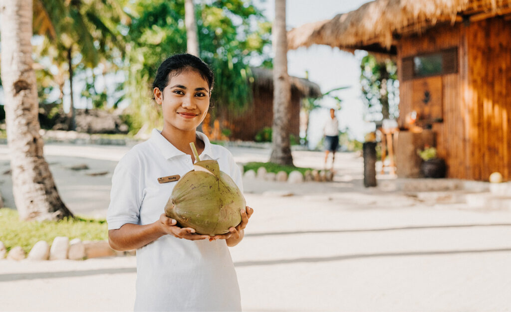 Member of the Spa Team at NIHI Sumba warmly welcoming guests to Nihioka Spa with a fresh coconut, standing in front of the spa’s bamboo reception area.