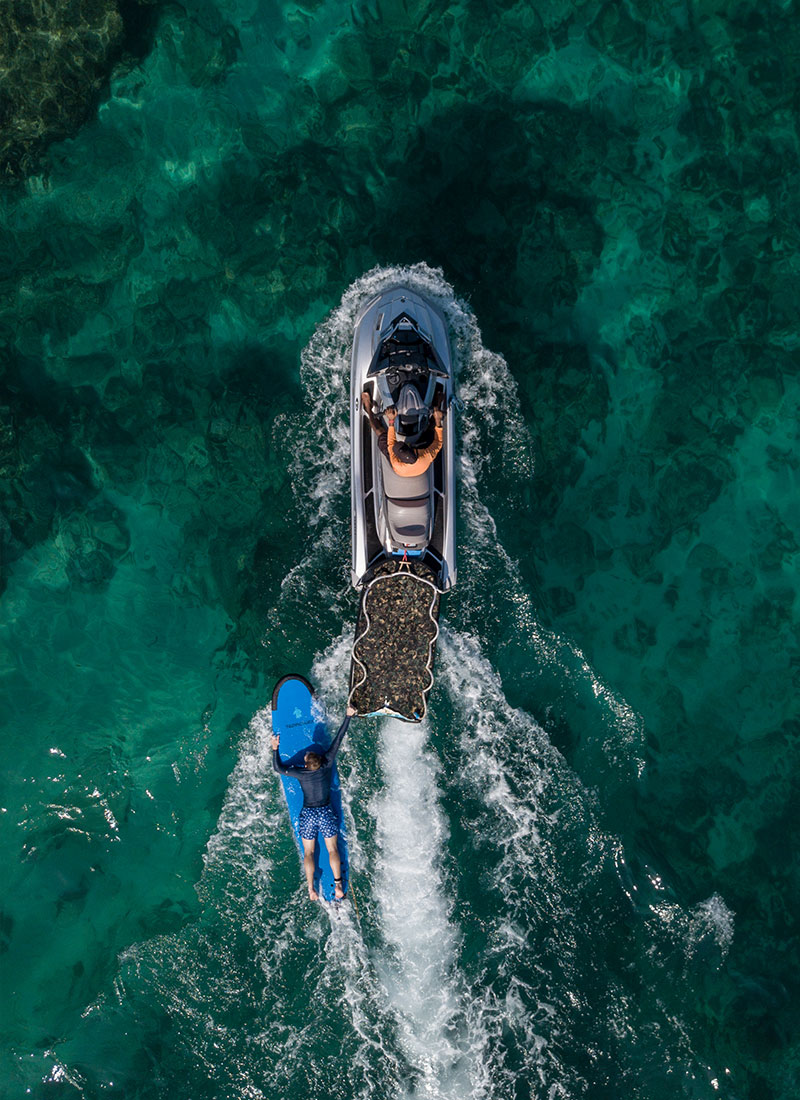 An aerial view of a jet ski towing a surfer over clear turquoise waters at NIHI® Sumba, showcasing the resort's water sports experiences in the Indian Ocean.