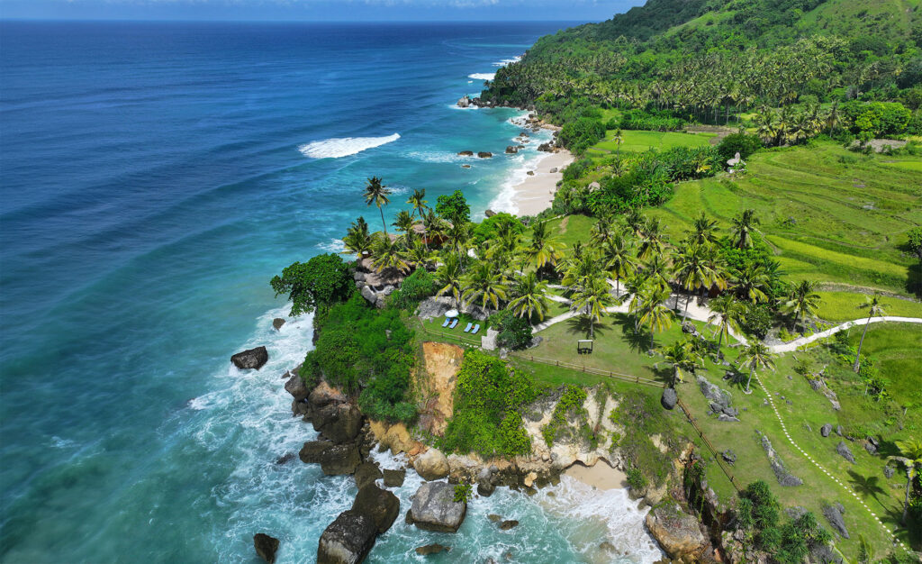 Aerial view of Nihioka Spa, nestled along the turquoise coastline of Sumba and surrounded by lush green fields and swaying palm trees—capturing the serene and secluded cliffside setting.