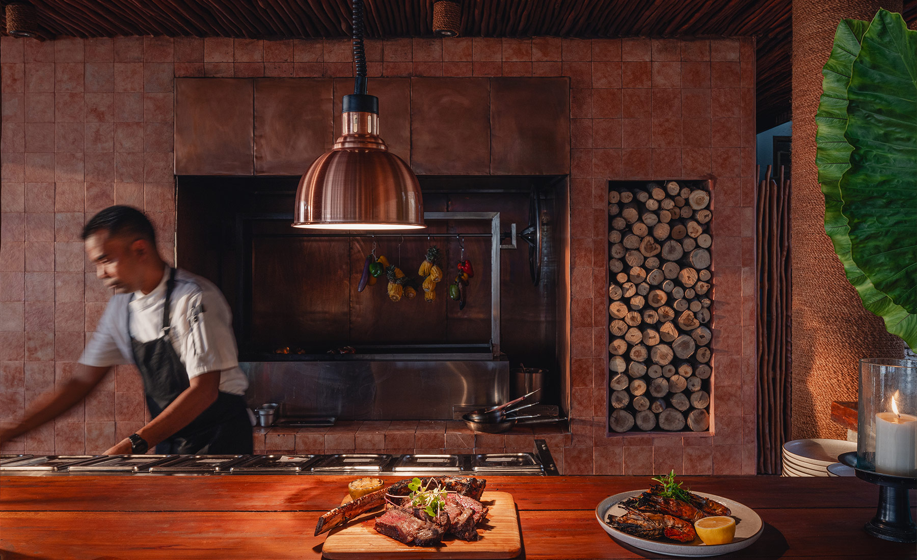 Chef in motion behind the counter at Ombak Restaurant in NIHI Sumba, preparing a gourmet dish under warm pendant lighting with grilled meat plated in the foreground and a rustic tiled backdrop.