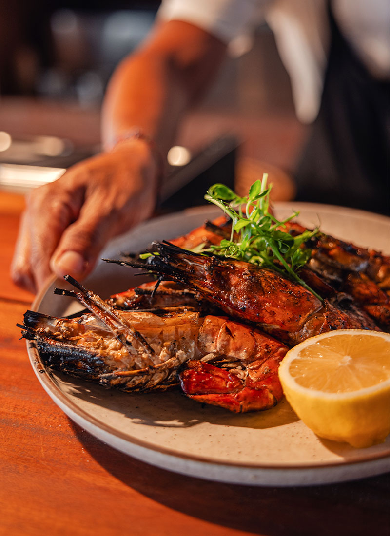 Close-up of grilled prawns garnished with fresh herbs and a slice of lemon, served as part of the seafood feast at Ombak Restaurant, NIHI Sumba. The dish showcases the fresh, locally sourced ingredients that are central to the resort's culinary offerings.