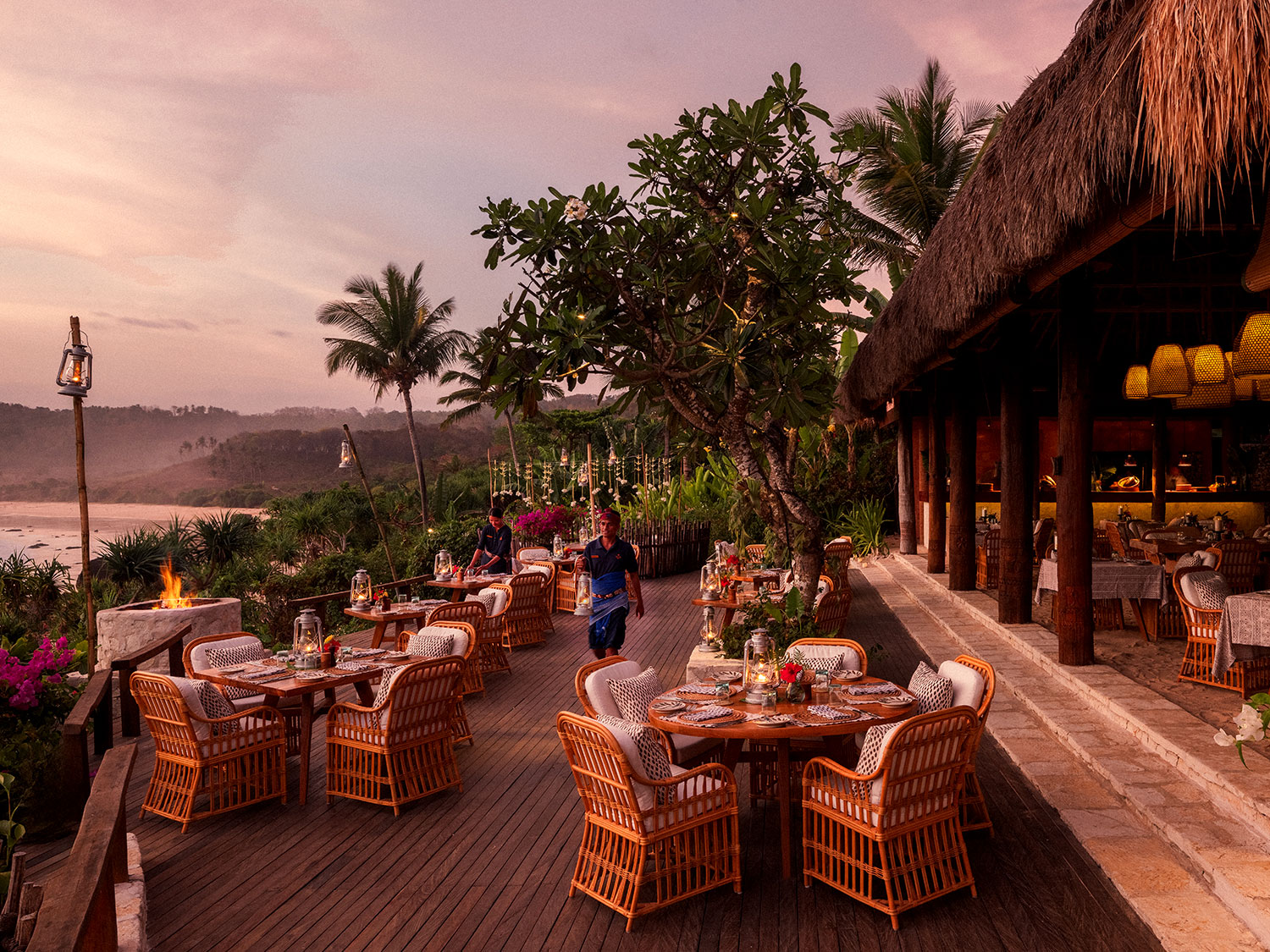 Outdoor dining setup at Ombak Restaurant, NIHI Sumba, during sunset, with elegant wicker tables set with plates and candles, surrounded by lush greenery and overlooking the 2.5km-long Nihiwatu Beach, as staff prepare the area to create a tranquil and romantic atmosphere.