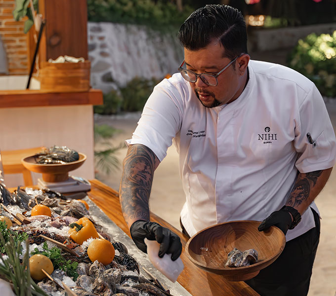 A chef at NIHI Sumba selecting fresh squid at the Seafood Market during the resort’s Friday seafood feast. Wearing a NIHI-branded uniform and gloves, he carefully picks from a variety of shellfish and seafood, preparing the display for an elegant beachfront dining experience.