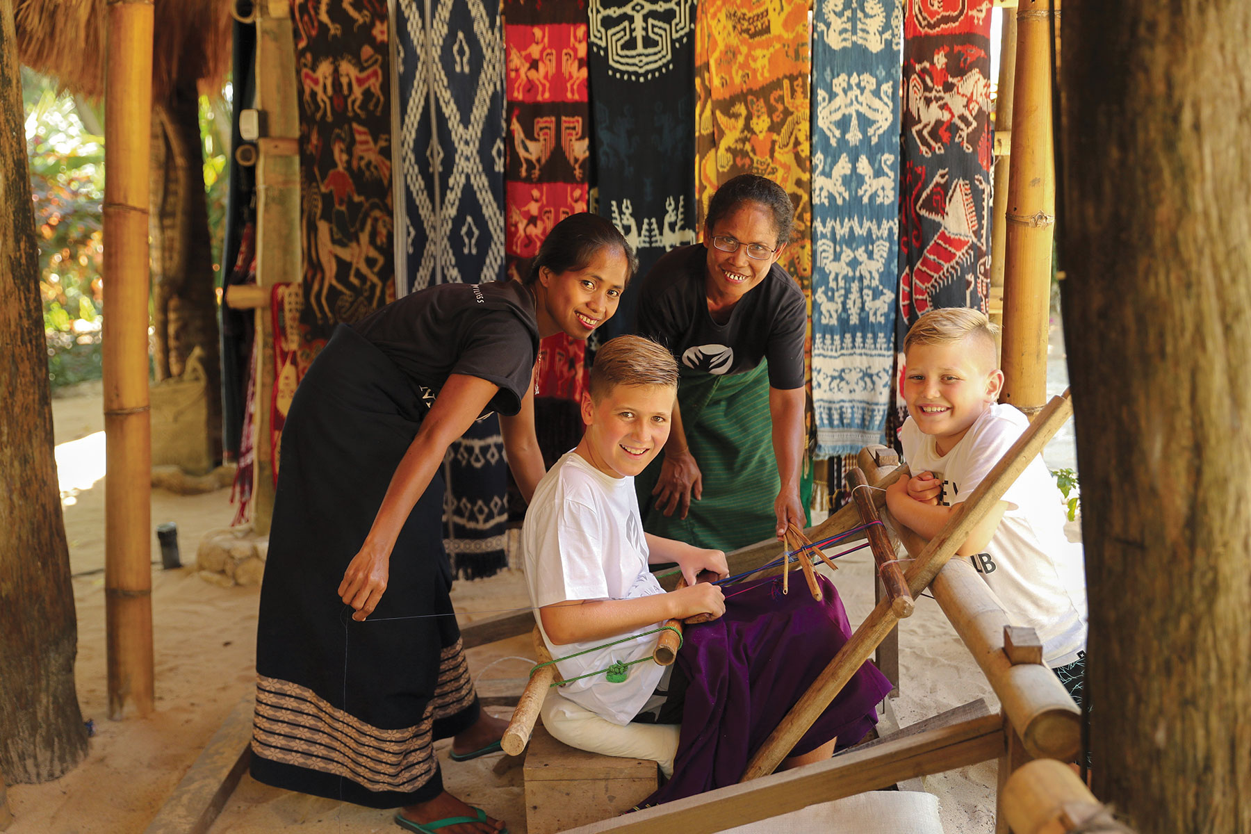Two young guests learning traditional tenun ikat weaving from Sumbanese artisans at NIHI Sumba, surrounded by vibrant handwoven textiles—an immersive cultural experience celebrating heritage and community craftsmanship.