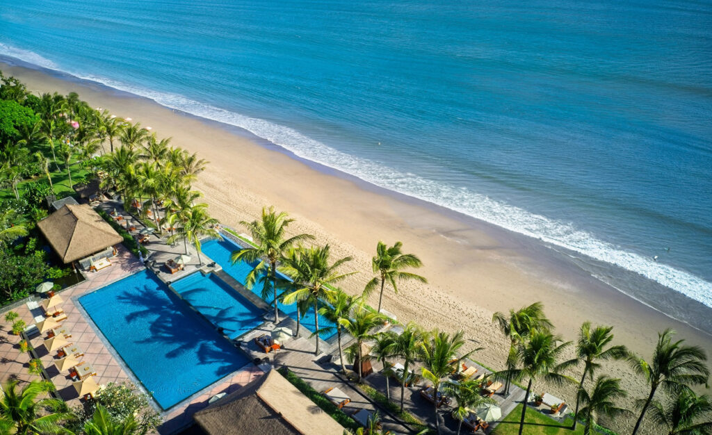 Aerial view of the pool at The Legian Seminyak, Bali—NIHI’s partner property—featuring a luxury beachfront setting with swaying palm trees, golden sands, and turquoise waters along Legian Beach.