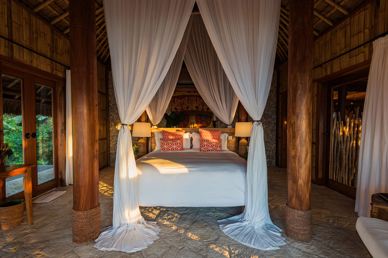 Interior view of Villa Rahasia's bedroom, showcasing a king-size bed with orange geometric pillows, framed by white sheer curtains. Traditional Sumbanese artwork adorns the wall behind the bed, complemented by soft lighting from bedside lamps, creating a romantic and serene ambiance.