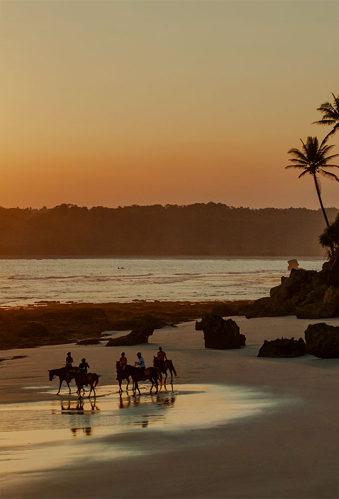 Guests on horseback riding along the shore at sunset during a festive Easter celebration on Nihiwatu’s 2.5 km beach, with giant colorful eggs nestled in the sand beneath swaying palms and golden skies.