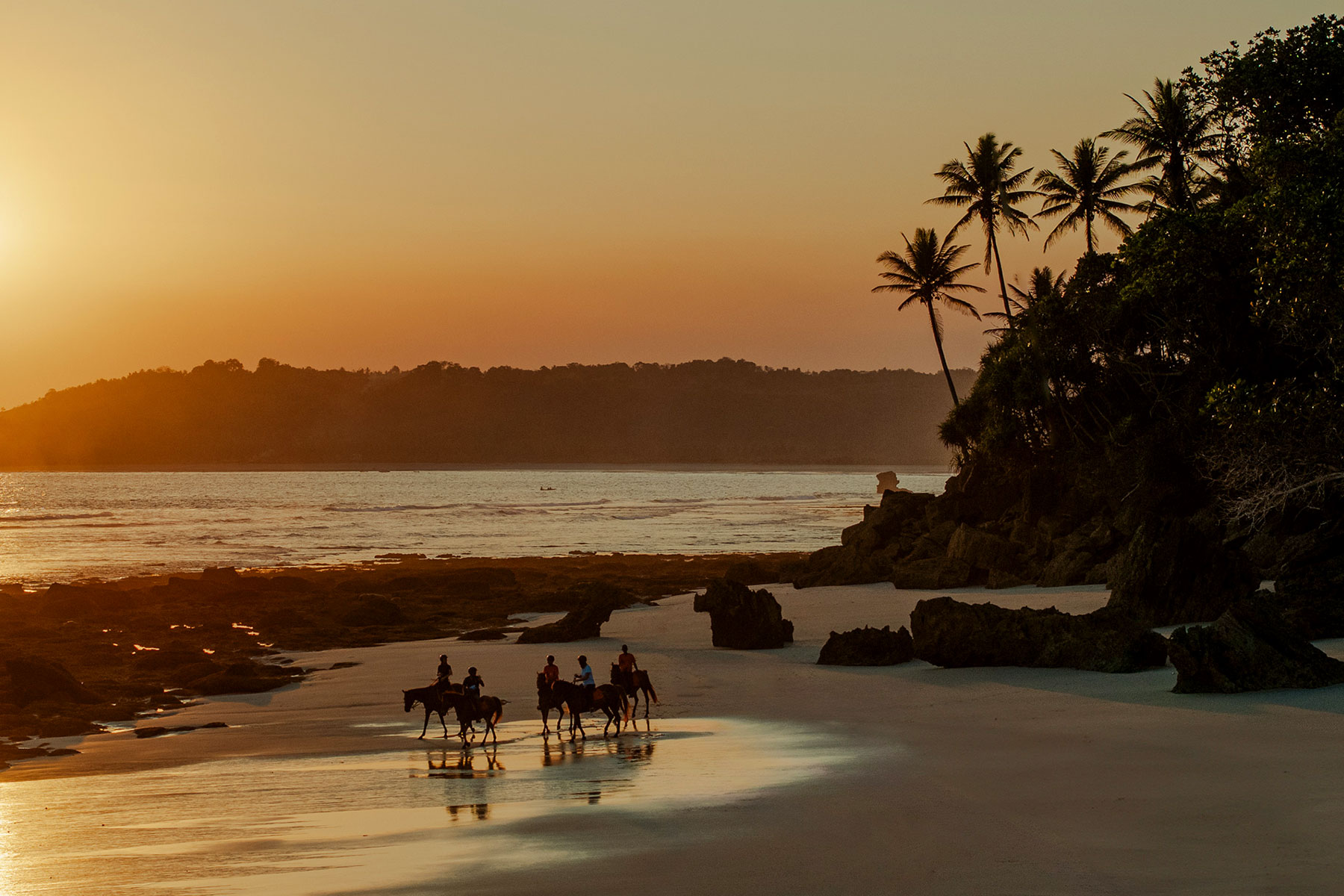 Guests on horseback riding along the shore at sunset during a festive Easter celebration on Nihiwatu’s 2.5 km beach, with giant colorful eggs nestled in the sand beneath swaying palms and golden skies.