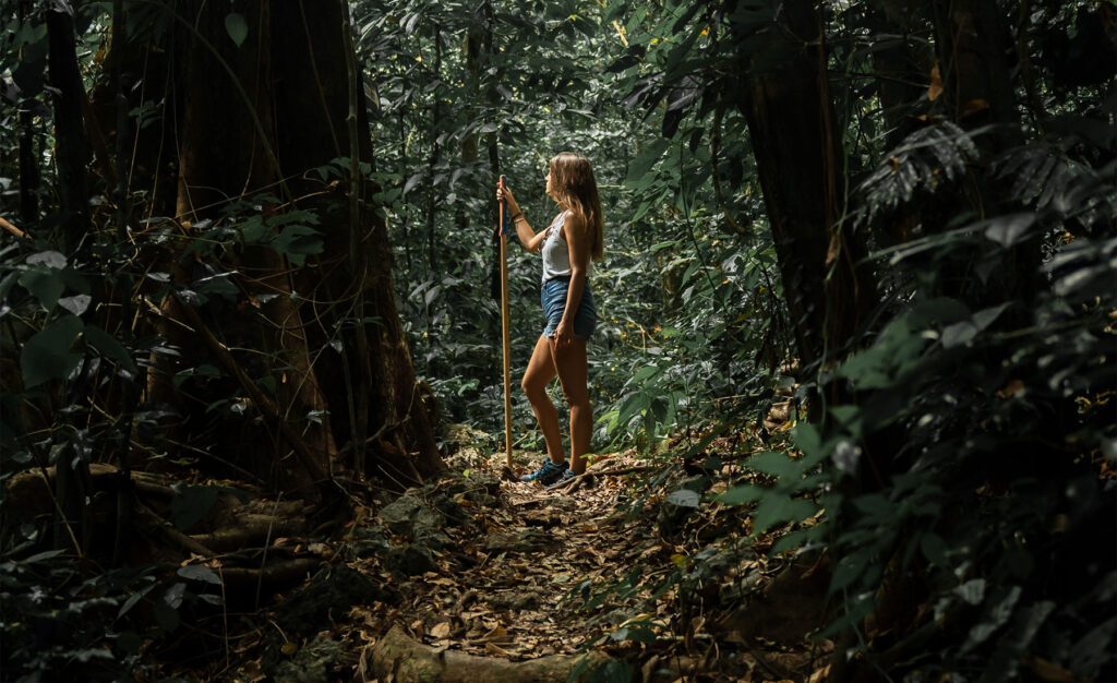 A female trekker explores the dense tropical forest on the Blue Waterfall Trek in Manupeu Tanah Daru National Park, surrounded by towering trees and lush jungle foliage.