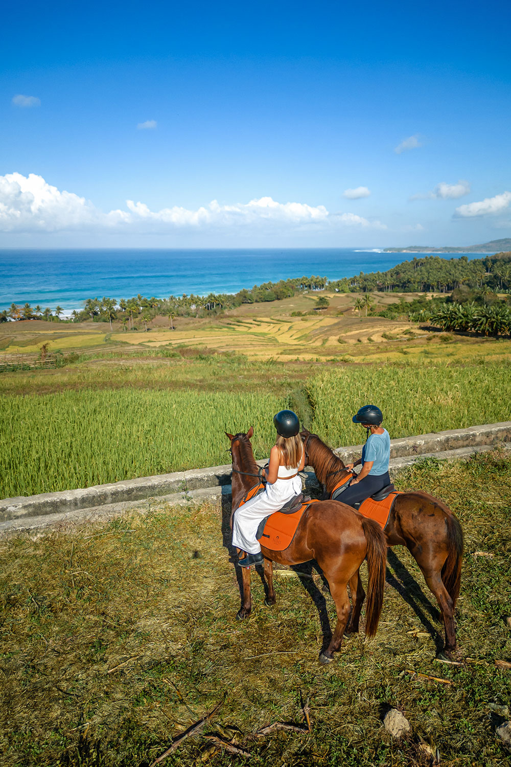 Two riders on horseback overlooking terraced rice fields and the Indian Ocean on a scenic trail ride experience at NIHI Sumba, blending coastal views with Sumba’s rural charm.