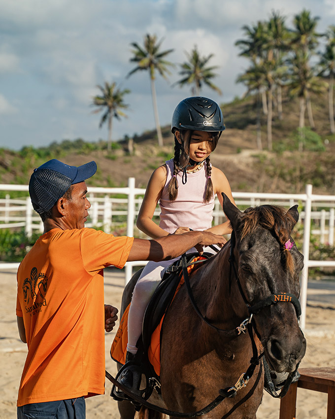 Sumba pony trainer guiding a young girl during a riding lesson at NIHI Sumba's Sandalwood Stables, with tropical scenery in the background.