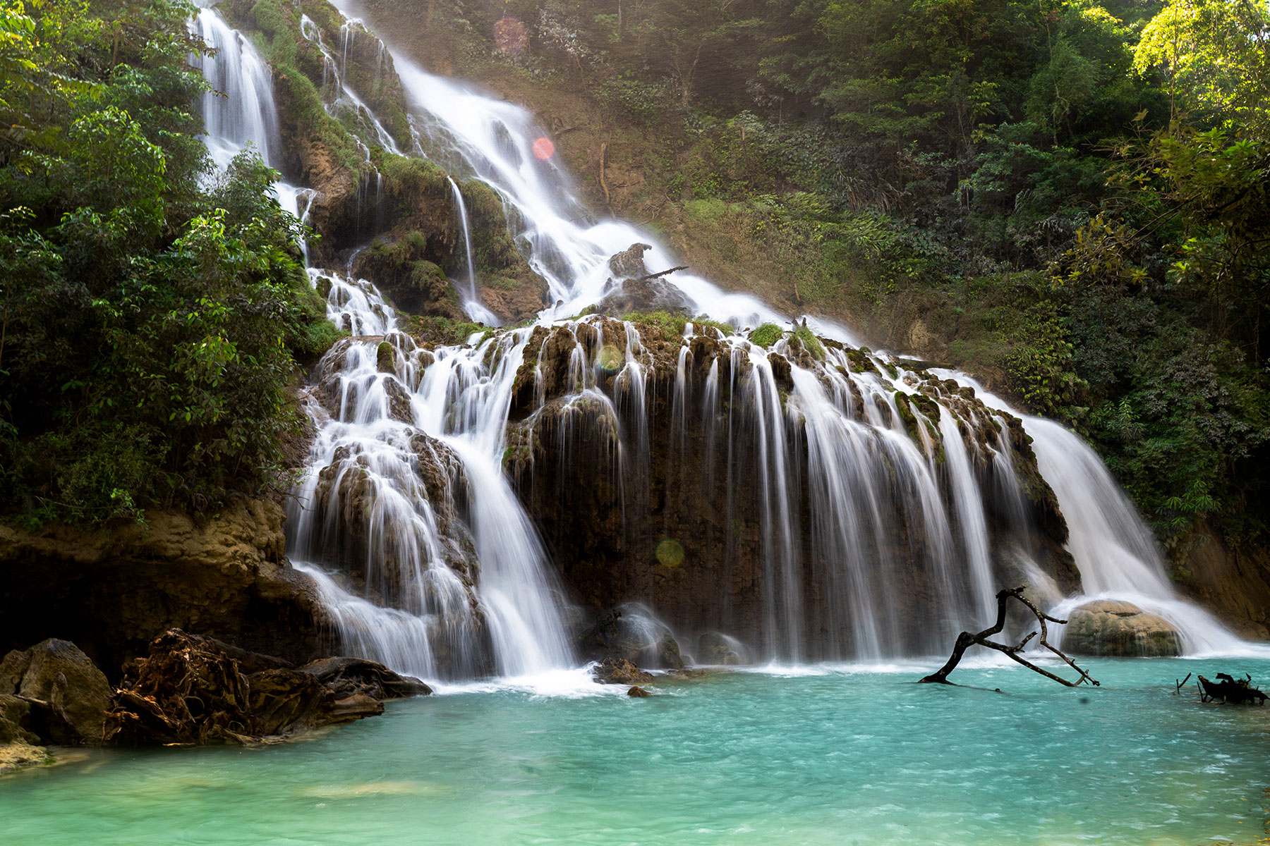 A breathtaking view of the Lapopu Waterfall in Sumba, with water cascading down over moss-covered rocks into a turquoise pool. A guest is seen swimming in the serene natural pool, surrounded by lush greenery and steep cliffs, making this location an ideal spot for relaxation and immersion in nature.
