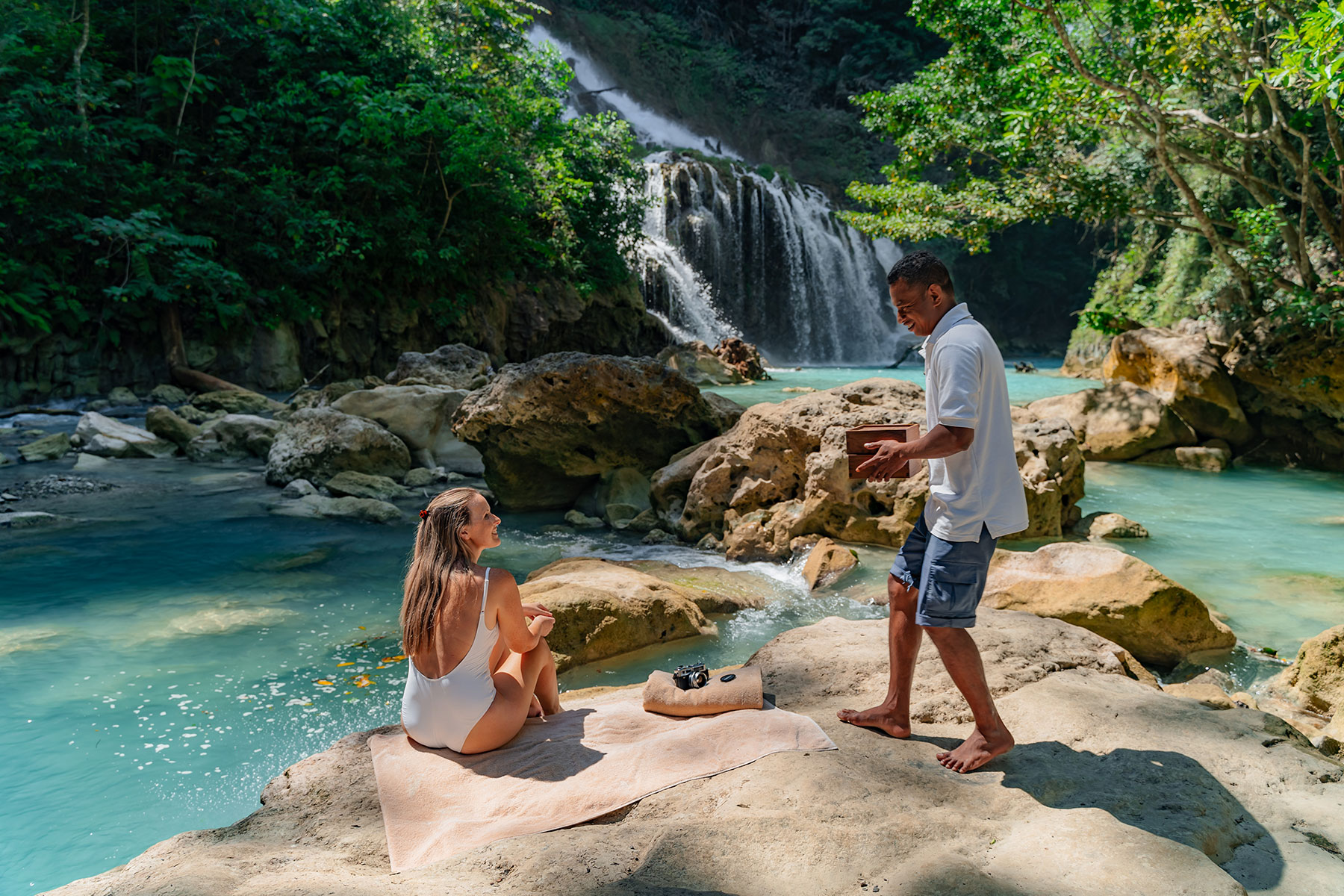 A guest relaxing on a rock at Lapopu Waterfall, surrounded by lush greenery and cascading water flowing into the natural pool below. A local guide stands nearby, carrying a picnic box, enhancing the experience with a thoughtful touch of hospitality.