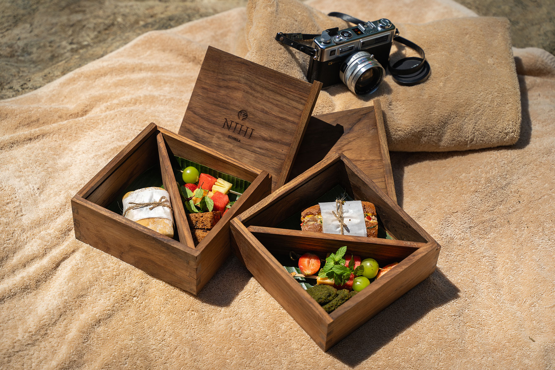 Elegant wooden lunch boxes with gourmet bites, fresh fruit, and artisanal sandwiches set on a soft towel beside a vintage camera, prepared for a NIHI picnic at Lapopu Waterfall.