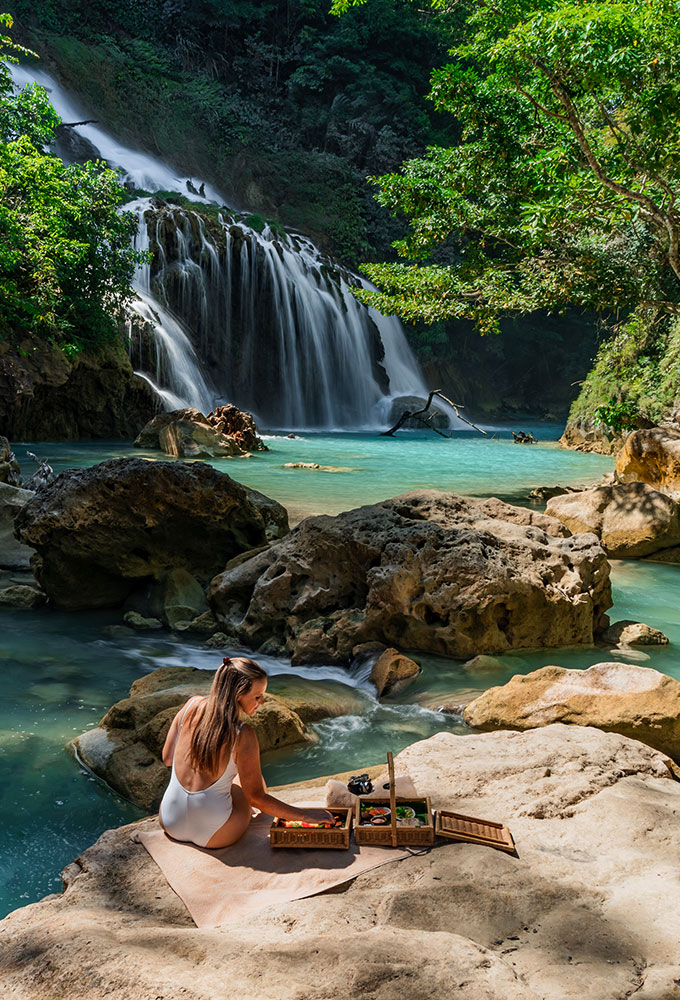 Woman enjoying a luxury riverside picnic near Lapopu Waterfall in Sumba, surrounded by lush jungle and turquoise water at NIHI resort.