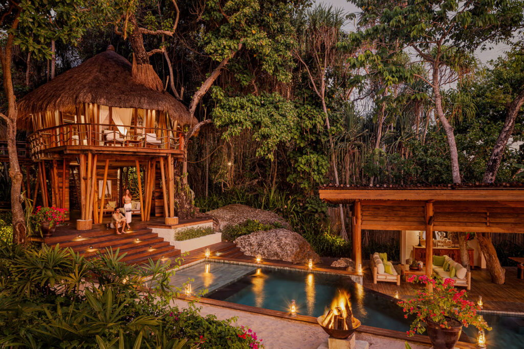 Mamole Treehouse at NIHI® Sumba, a bamboo villa with open-air lounge spaces surrounding a private pool illuminated by lanterns and a fire pit, nestled in a lush forest setting.