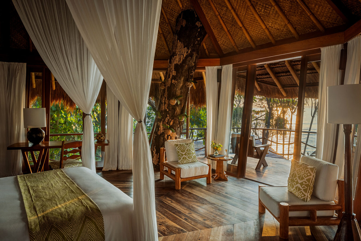 Interior of a Mamole Treehouse Villa suite at NIHI Sumba, featuring a canopy king bed, natural wood furnishings, and a striking tree trunk integrated into the room’s design, with floor-to-ceiling glass doors opening to a private deck with ocean and jungle views.
