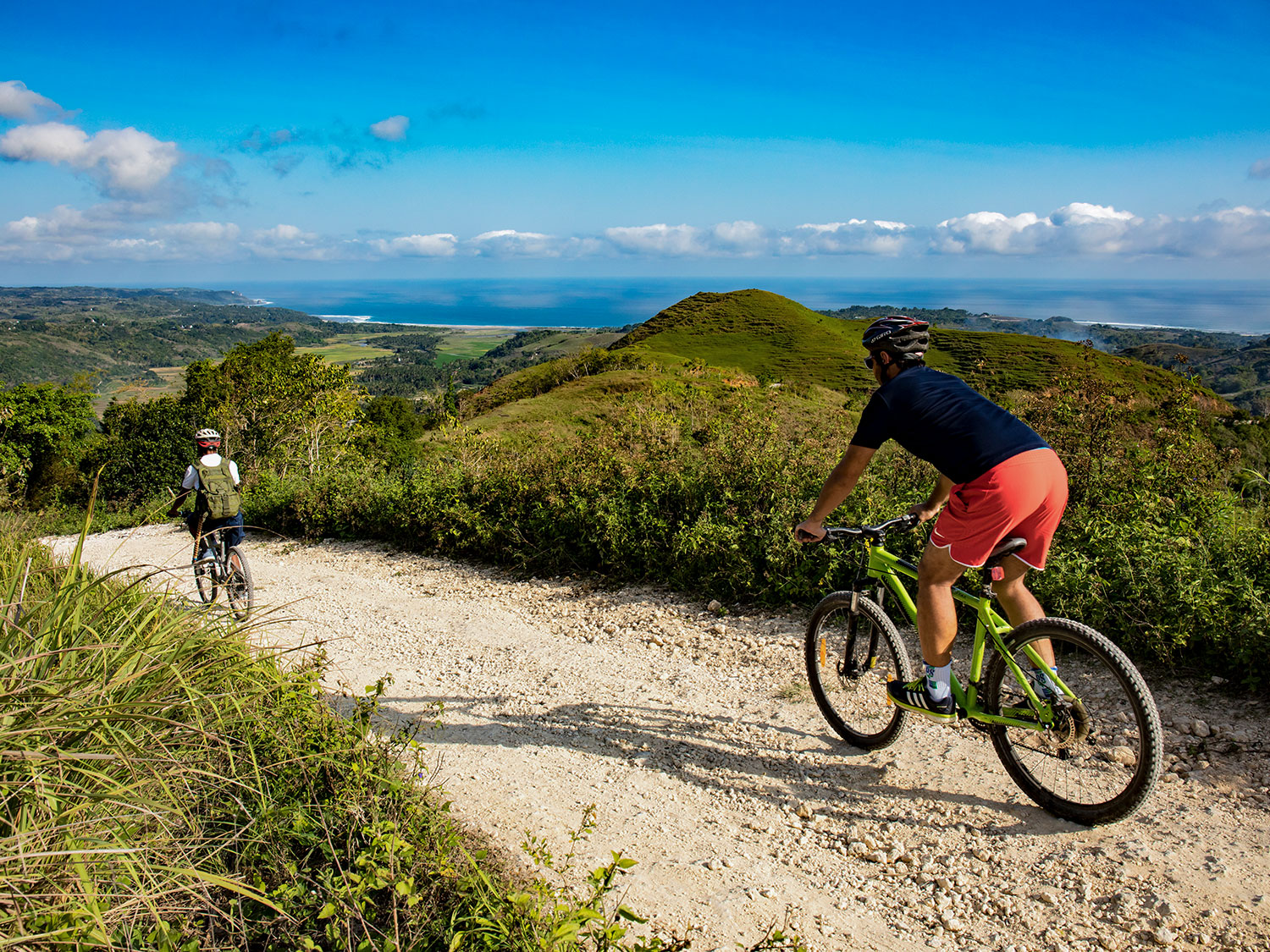 Two guests mountain biking along a winding gravel trail atop the lush hills of Sumba Island, with panoramic views of the countryside and ocean horizon.