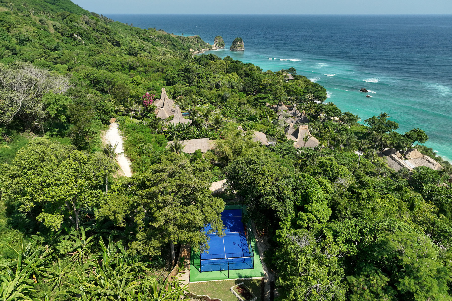Aerial view of NIHI Sumba’s vibrant padel court set amid lush tropical gardens, with guests playing a match against the backdrop of thatched villas and the Indian Ocean — a highlight of the resort’s Active Wellness program.