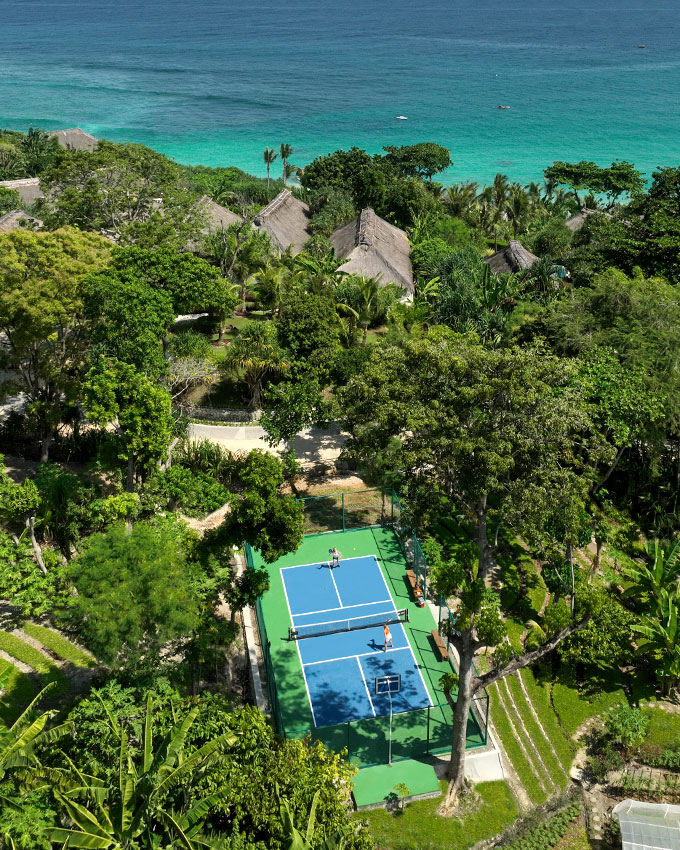 Aerial view of the Pickleball Court at NIHI Sumba, surrounded by lush jungle and overlooking the crystal-clear ocean. The vibrant blue court sits harmoniously within the tropical landscape, offering a serene and scenic setting for pickleball enthusiasts.