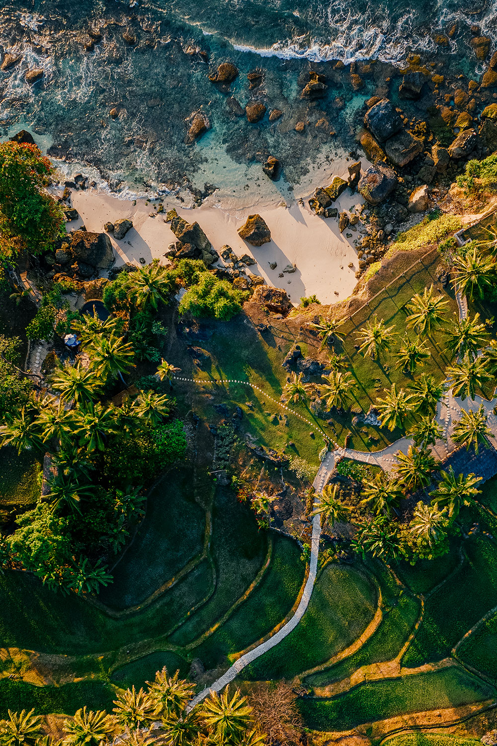 Aerial view of the Nihioka Spa rice terraces at NIHI® Sumba, with lush green paddies cascading down the valley slopes toward the Indian Ocean, surrounded by tropical foliage and stone-lined garden paths.