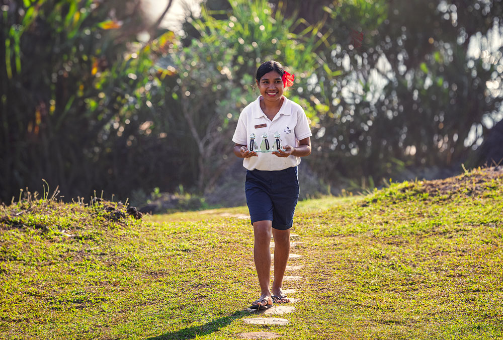 A smiling spa therapist at Nihioka Spa walks along a lush garden path, carrying a tray of essential oils for treatments, surrounded by tropical greenery and illuminated by warm afternoon light.