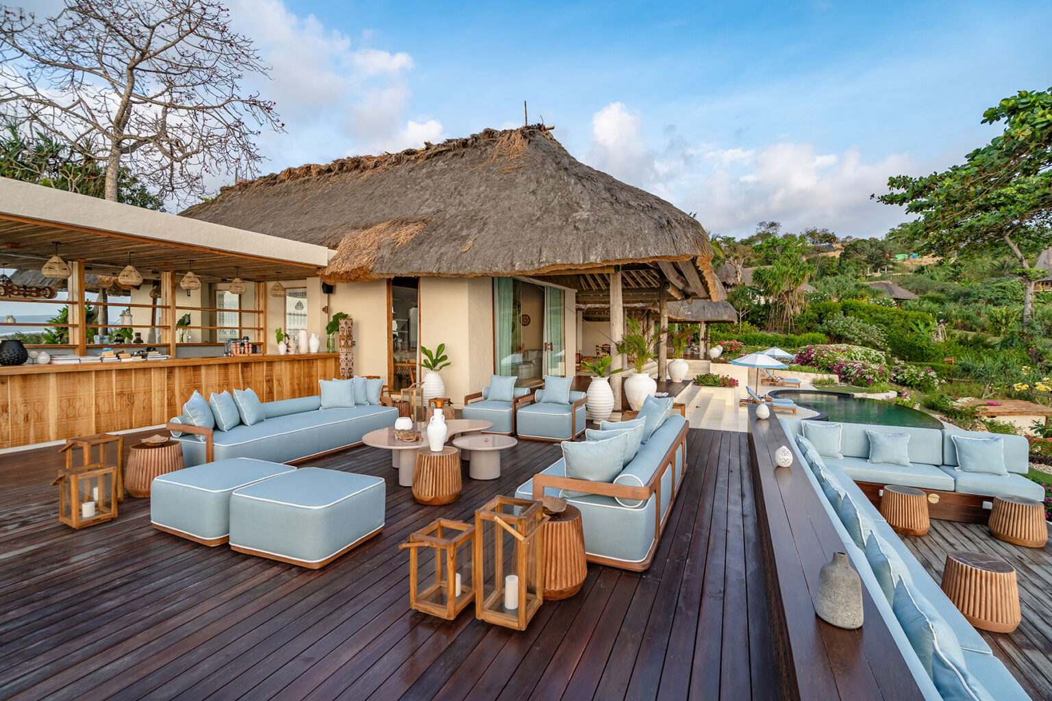 Outdoor lounge area of Putri Kasambi villa, showcasing light blue cushioned seating, thatched roof architecture, a private bar, and a curved pool surrounded by vibrant gardens at NIHI Sumba.