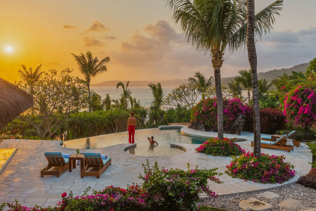 Sunset view from the infinity-edge pool at Putri Wamoro Beach Villa in NIHI Sumba, with two guests enjoying the tranquil atmosphere surrounded by lush greenery and overlooking the ocean horizon.