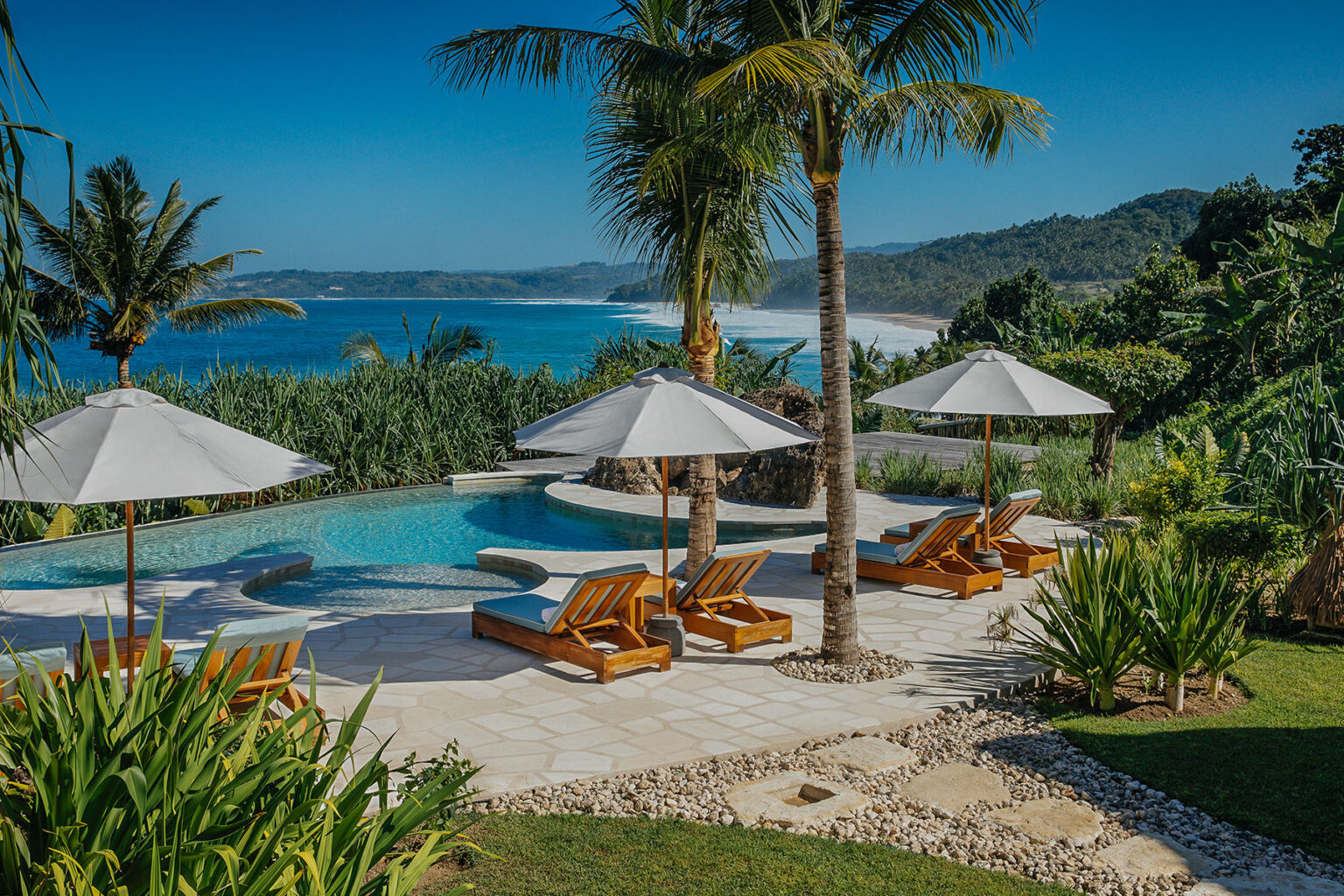 Panoramic view of the private pool area at Putri Wamoro Beach Villa in NIHI Sumba, featuring wooden sun loungers with white umbrellas, swaying palm trees, and a stunning backdrop of the ocean and rugged coastline.