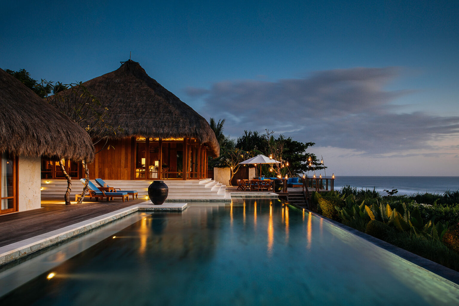 Twilight view of Raja Mandaka’s main villa and infinity pool overlooking the Indian Ocean, with ambient lighting and thatched-roof architecture at NIHI Sumba.