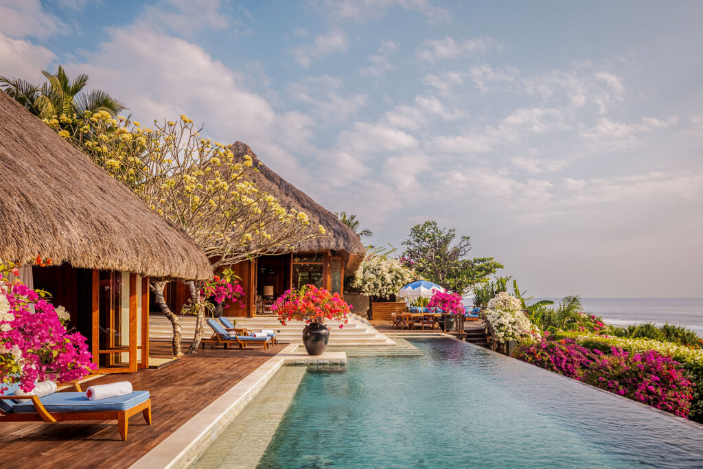 Main pool of Raja Mandaka, the Owner’s Villa within the nine-bedroom Mandaka Estate at NIHI® Sumba, surrounded by blooming tropical flowers and traditional thatched-roof architecture.