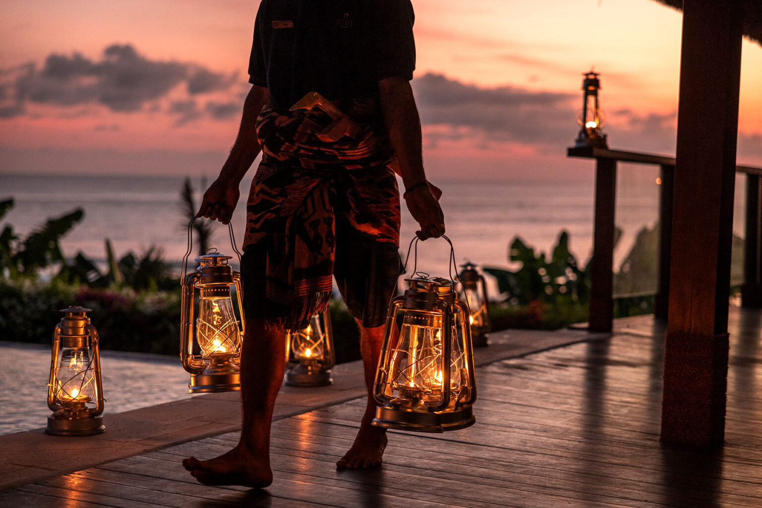 Barefoot NIHI Sumba staff member carrying vintage lanterns at sunset, creating a warm and intimate ambiance overlooking the ocean.
