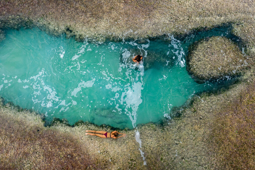 Two guests enjoying the clear turquoise water of a tidal rock pool at Cleopatra Rock during low tide, with one guest swimming and the other relaxing by the pool's edge at Nihiwatu Beach.