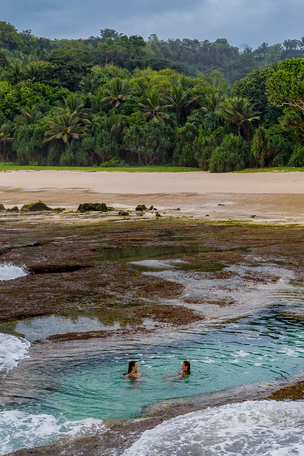 Two guests enjoying a swim in a tidal rock pool at Nihiwatu Beach, with the lush tropical forest in the background and the expansive beach just beyond, surrounded by natural coral formations.
