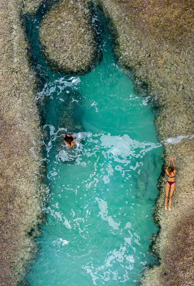 Two guests enjoying the clear turquoise water of a tidal rock pool at Cleopatra Rock during low tide, with one guest swimming and the other relaxing by the pool's edge at Nihiwatu Beach.