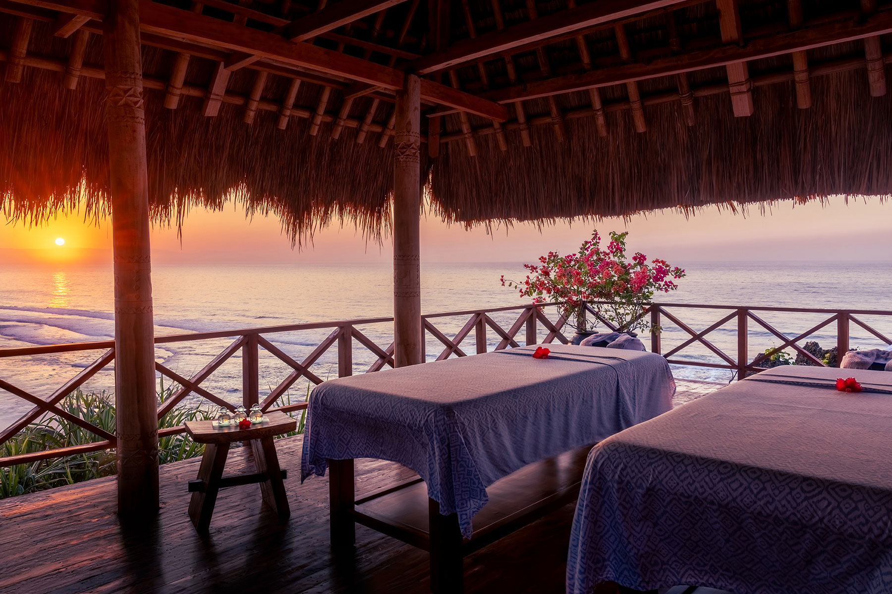 Two massage tables inside an open-air thatched-roof treatment bale at NIHI® Sumba’s Spa Safari™, overlooking the Indian Ocean at sunrise, with warm golden light streaming through the roof and soft waves below.
