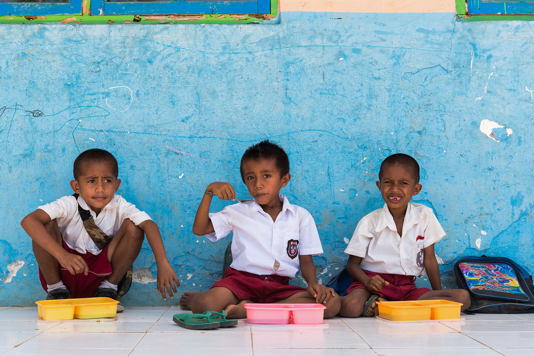 Three young Sumbanese boys in school uniforms enjoying a midday meal from colorful lunchboxes, seated against a bright blue classroom wall—part of the school nourishment initiatives by the Sumba Foundation, Burch Family Foundation and NIHI Sumba.