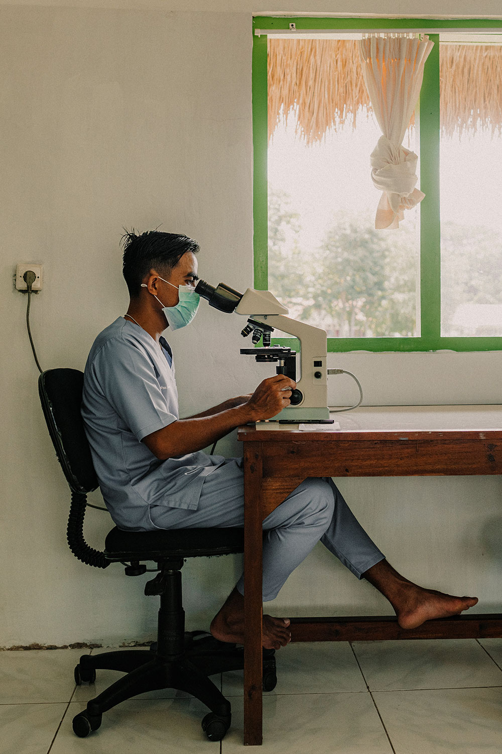 A healthcare worker in scrubs and a face mask examines samples under a microscope at a Sumba Foundation clinic, highlighting the foundation’s ongoing efforts in malaria prevention and community health at NIHI Sumba.
